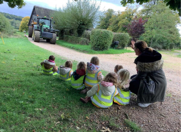 A group of nursery children and their wave to a tractor. The children are in high-vis jackets and safe with their teacher.