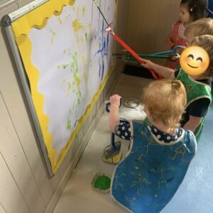 A group of toddlers paint on a vertically mounted piece of paper.