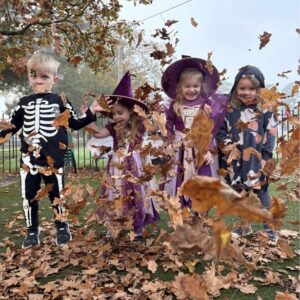 A group of toddlers in Halloween costumes kick up leaves in a flurry of excitement.